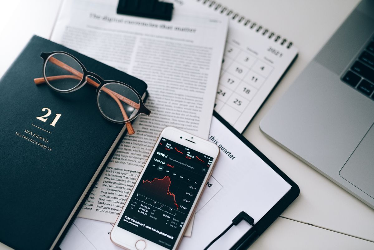 A research desk with documents, eyeglasses, a smartphone showing a market chart and a laptop — the working surface on which editorial articles about AI and currency exposure are drafted.